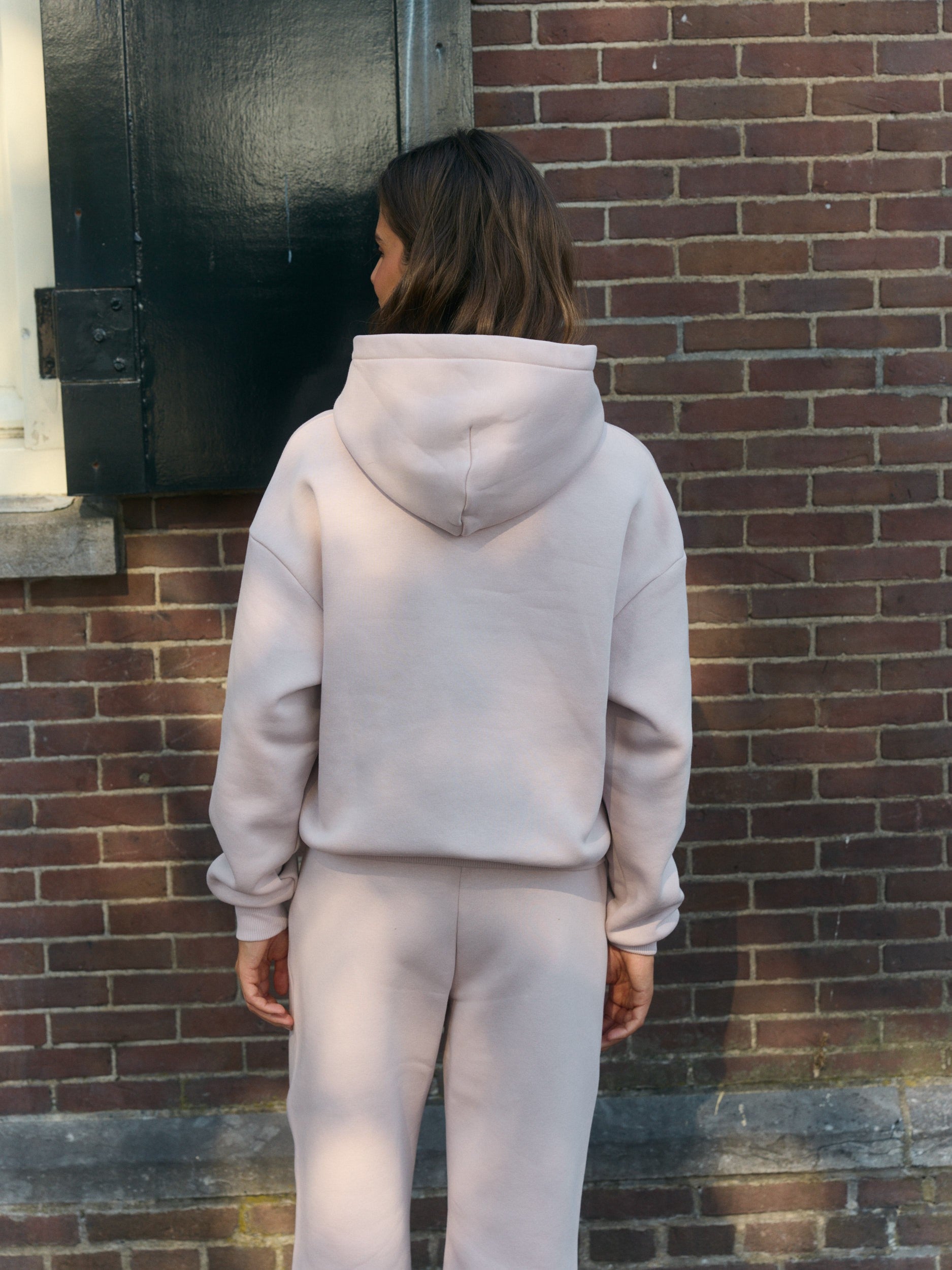 Back view of a woman wearing a dusty pink hoodie and matching sweatpants against a brick wall.